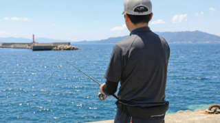 夏の堤防で腰巻き救命具を着用しUVカットキャップを被って釣りをする人物、強い日差しの中の日中シーン