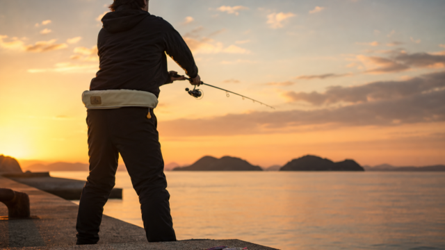 夕まずめの瀬戸内海の堤防でエギングロッドを構える釣り人。足元にはエギがいくつか並び、遠くに島影が見える