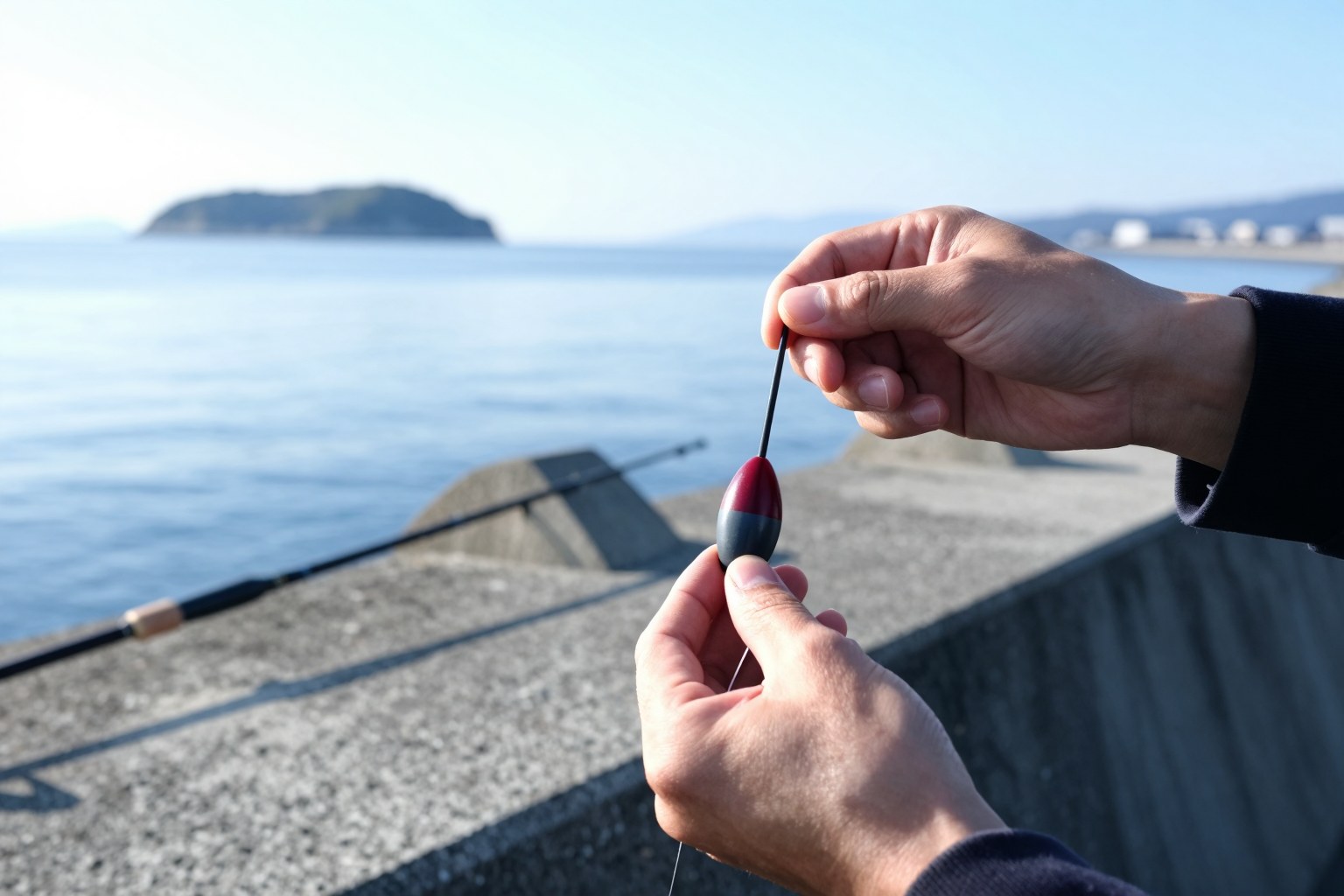 朝の堤防で、円錐ウキを通した道糸にウキ止めを結ぶ釣り人の手元。奥に磯竿と海