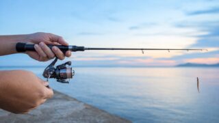 明るい夕暮れの堤防で、メバリングロッドとスピニングリールを構える両手の手元アップ。穂先からジグヘッドとワームが垂れる