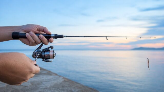 明るい夕暮れの堤防で、メバリングロッドとスピニングリールを構える両手の手元アップ。穂先からジグヘッドとワームが垂れる