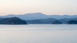朝の瀬戸内海を水面近くの低い視点から見た風景。重なる島影と、沖に小さく浮かぶ遊漁船
