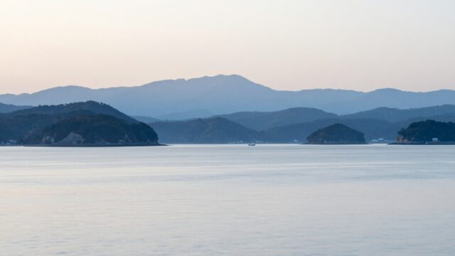 朝の瀬戸内海を水面近くの低い視点から見た風景。重なる島影と、沖に小さく浮かぶ遊漁船