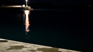 夜の瀬戸内の漁港。常夜灯の光が岸壁際の水面に落ち、明るい部分と暗い部分の境目ができている様子を、水面すれすれのローアングルから寄りで写した写真