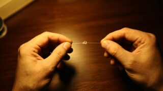 出船前夜の作業机で、デスクライトの下、細い釣り糸の結び目を両手の指先で締め込んでいる手元を、真上から寄って写した写真