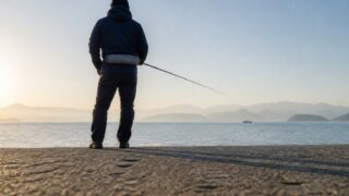 朝の江田島の護岸で、腰に救命具を着けた釣り人が瀬戸内の海に向かって竿を出している後ろ姿を、足元のコンクリート面に近いローアングルから中景で写した写真