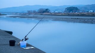 朝の河口。砂泥が混じった浅い水際に短い投げ竿を立てかけ、足元に小さなバケツと仕掛けの小袋を置いた様子を、水面と同じ高さから中景で写した写真